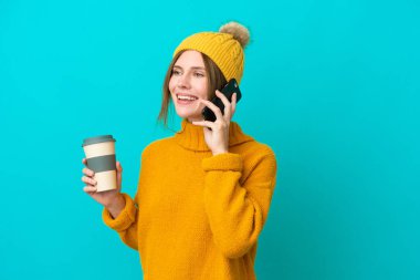 Kışlık ceket giyen genç İngiliz kadın mavi arka planda izole bir şekilde kahve ve bir cep telefonu tutuyor.