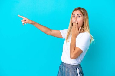 Sarışın Uruguaylı kız mavi arka planda izole edilmiş sürpriz bir ifadeyle işaret ederken