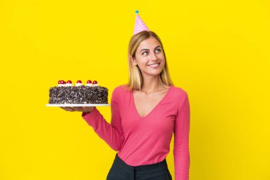 Blonde Uruguayan girl holding birthday cake isolated on yellow background thinking an idea while looking up