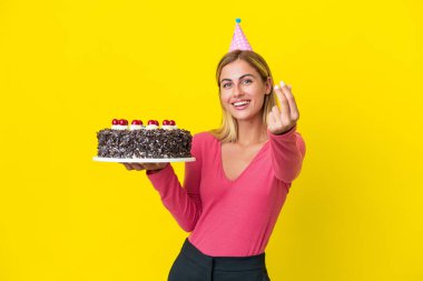 Blonde Uruguayan girl holding birthday cake isolated on yellow background making money gesture