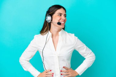 Tele pazarlamacı İtalyan kadın mavi arka planda izole edilmiş bir kulaklıkla çalışıyor. Kolları kalçasında ve gülümsüyor.