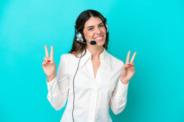 Tele pazarlamacı İtalyan kadın mavi arka planda izole edilmiş bir kulaklıkla çalışıyor. İki eliyle zafer işareti gösteriyor.