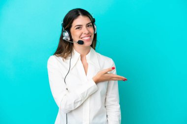 Tele pazarlamacı İtalyan kadın mavi arka planda izole edilmiş bir kulaklıkla çalışırken bir fikir sunuyor ve gülümsüyor.