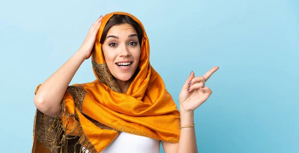 若いですモロッコの女性とともに伝統的な衣装青の背景に隔離された驚きと側面に指を指す — ストック写真