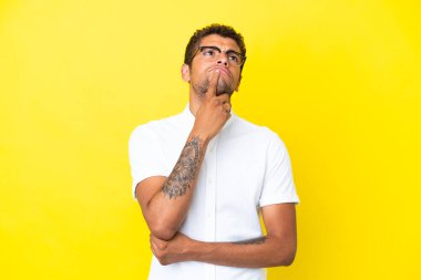 Genç ve yakışıklı Brezilyalı bir adam sarı arka planda izole edilmiş, yukarı bakarken şüpheleri var.