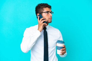 Genç Brezilyalı iş adamı mavi arka planda izole edilmiş. Elinde kahve ve cep telefonu var.