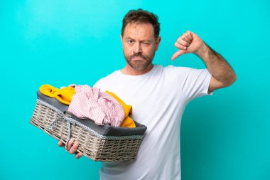 Mavi arka planda izole edilmiş çamaşır sepetini tutan orta yaşlı bir kadın negatif ifadeli baş parmağını gösteriyor.