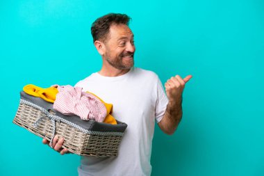 Orta yaşlı bir kadın mavi arka planda izole edilmiş bir çamaşır sepetini elinde tutuyor. Ürünü sunmak için yan tarafa bakıyor.