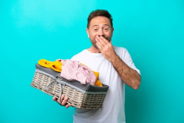Mavi arka planda bir çamaşır sepeti tutan orta yaşlı bir kadın mutlu ve gülümseyen bir ağızla elini kapatıyor.