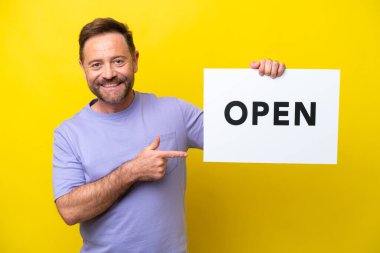 Orta yaşlı, beyaz bir adam sarı arka planda izole edilmiş. Elinde açık yazılı bir pankart var.