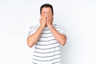 Beyaz arka planda izole edilmiş beyaz tenli genç adam gözlerini elleriyle kapatıyor.
