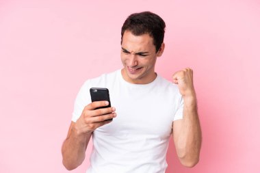 Young caucasian man isolated on pink background using mobile phone and doing victory gesture