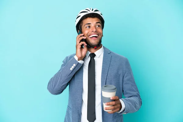 Young business Brazilian man with bike helmet isolated on blue background holding coffee to take away and a mobile