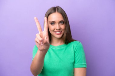 Mor arka planda izole edilmiş genç beyaz kadın gülümsüyor ve zafer işareti gösteriyor.