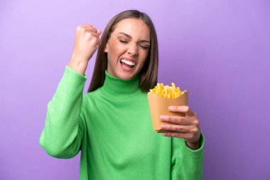 Young caucasian woman holding fried chips on purple background celebrating a victory