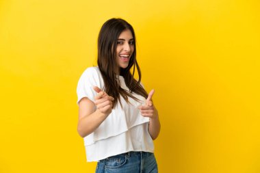 Sarı arka planda izole edilmiş genç beyaz kadın ön tarafı işaret ediyor ve gülümsüyor.
