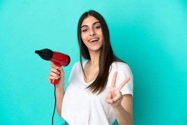 Mavi arka planda izole edilmiş saç kurutma makinesini tutan beyaz bir kadın gülümsüyor ve zafer işareti gösteriyor.