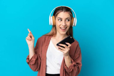 Genç beyaz kadın mavi arka planda izole edilmiş. Cep telefonuyla müzik dinliyor ve şarkı söylüyor.