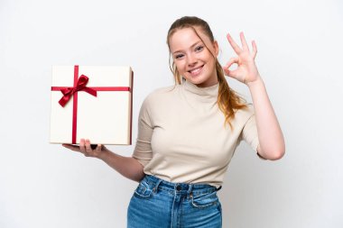 Beyaz arka planda izole edilmiş bir hediye tutan genç beyaz kadın parmaklarıyla tamam işareti gösteriyor.