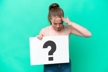 Young caucasian woman isolated on green background holding a placard with question mark symbol with surprised expression