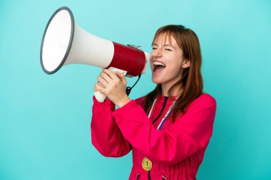 Redhead girl with medals isolated o blue background shouting through a megaphone