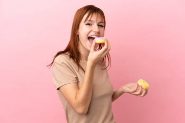Kızıl saçlı kız pembe arka planda izole edilmiş donut yiyor.