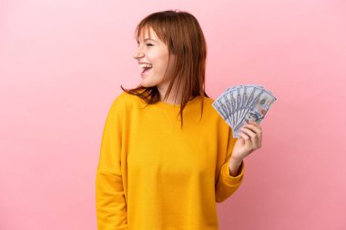 Kızıl saçlı kız pembe arka planda izole edilmiş bir sürü para alıyor ve yan yana gülüyor.