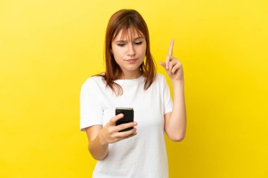 Kızıl saçlı kız sarı arka planda cep telefonu ve parmak kaldırarak izole edilmiş.