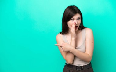 Yeşil arka planda izole edilmiş genç Rus kadın bir ürün sunmak ve bir şeyler fısıldamak için kenarı işaret ediyor.