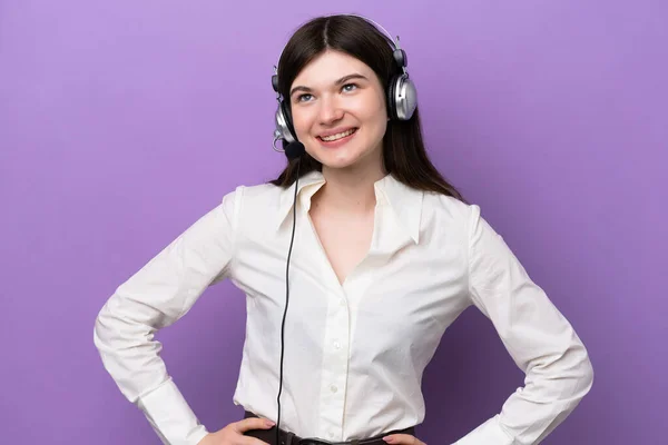 Telemarketer Russian woman working with a headset isolated on purple background posing with arms at hip and smiling