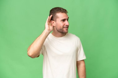 Genç beyaz adam yeşil krom arka planda izole edilmiş bir şekilde elini kulağına koyarak bir şeyler dinliyor.