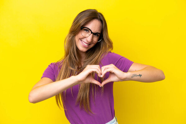 Молодая белая женщина изолирована на желтом фоне с очками, создающими сердце руками