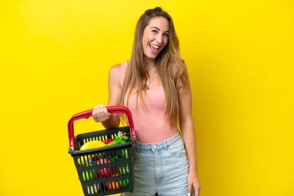 黄色の背景に孤立した食べ物でいっぱいの買い物かごを持っている若い白人女性と驚きの顔の表情 — ストック写真
