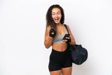 Spor çantalı genç Arap kadın, beyaz arka planda izole edilmiş ve telefon zafer pozisyonunda.