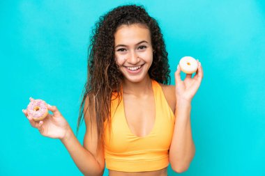 Mavi arka planda izole edilmiş genç bir Arap kadın elinde mutlu bir ifadeyle donut tutuyor.