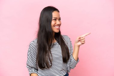 Pembe arka planda izole edilmiş genç Kolombiyalı bir kadın parmak göstererek bir ürün sunuyor.