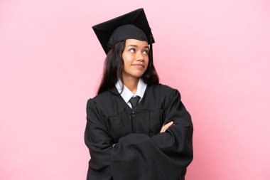 Üniversiteden mezun genç bir Kolombiyalı kadın pembe arka planda izole edilmiş. Omuzlarını kaldırırken şüpheli hareketler yapıyor.