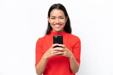 Beyaz arka planda izole edilmiş genç Kolombiyalı kadın kameraya bakıyor ve cep telefonunu kullanarak gülümsüyor.