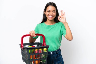 Genç Kolombiyalı bir kadın elinde yiyecek dolu bir sepet tutuyor. Beyaz arka planda izole edilmiş. Parmaklarıyla tamam işareti gösteriyor.