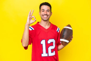 İzole edilmiş sarı arka planda ragbi oynayan genç yakışıklı adam parmaklarıyla işaret veriyor.