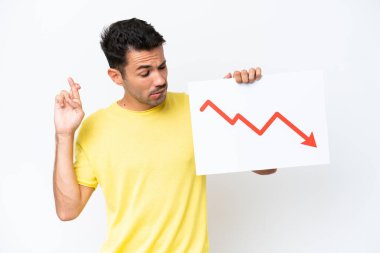 İzole edilmiş beyaz arkaplan üzerinde genç yakışıklı bir adam azalan bir istatistik ok sembolü ve şans dileyen bir işaret tutuyor.