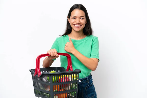 若いコロンビア人女性が白い背景に誇りと自己満足的に隔離された食べ物でいっぱいのショッピングバスケットを持っている — ストック写真