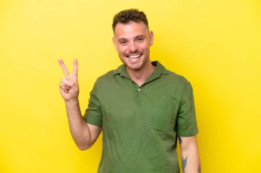 Sarı arka planda izole edilmiş yakışıklı genç adam gülümsüyor ve zafer işareti gösteriyor.