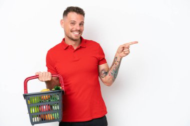 Beyaz arka planda izole edilmiş yiyecek dolu bir alışveriş sepeti tutan beyaz bir adam var.