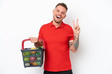 Beyaz arka planda izole edilmiş yiyecek dolu bir sepet taşıyan beyaz bir adam gülümsüyor ve zafer işareti gösteriyor.