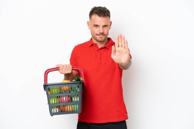 Beyaz arka planda izole edilmiş yiyecek dolu bir alışveriş sepeti tutan beyaz bir adam dur hareketi yapıyor.