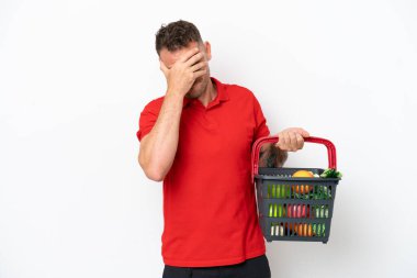 Beyaz arka planda, yorgun ve hasta bir ifadeyle izole edilmiş yiyecek dolu bir alışveriş sepeti tutan beyaz bir adam.