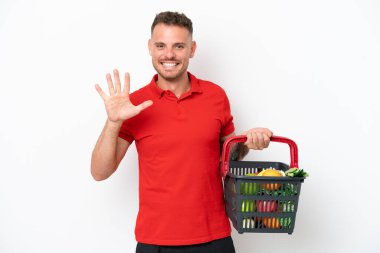 Beyaz arka planda izole edilmiş yiyecek dolu bir alışveriş sepeti tutan beyaz bir adam beş parmağıyla sayıyor.