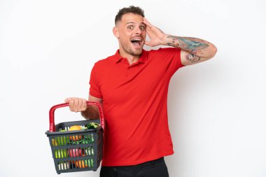 Beyaz arka planda, sürpriz bir ifadeyle izole edilmiş yiyecek dolu bir alışveriş sepeti tutan genç beyaz bir adam.