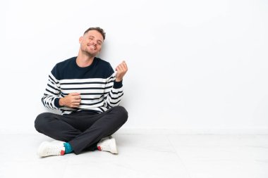 Yerde oturan genç adam, beyaz arka planda tek başına gitar hareketi yapıyor.